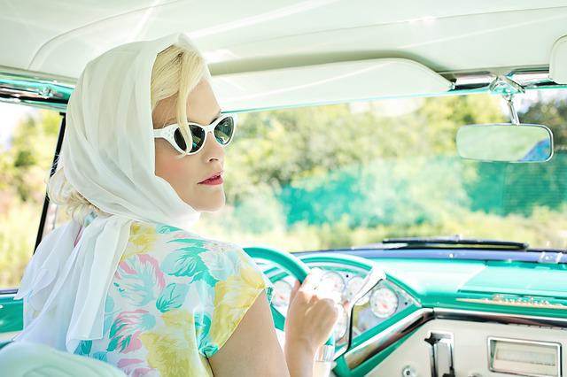 Kvinna klädd i klassiska 50-talskläder och solglasögon, bakom ratten i en gammal amerikanare (bil).